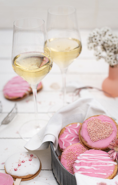 Zwei Gläser Sekt mit rosa verzierten Keksen auf einer weißen Tafel.
